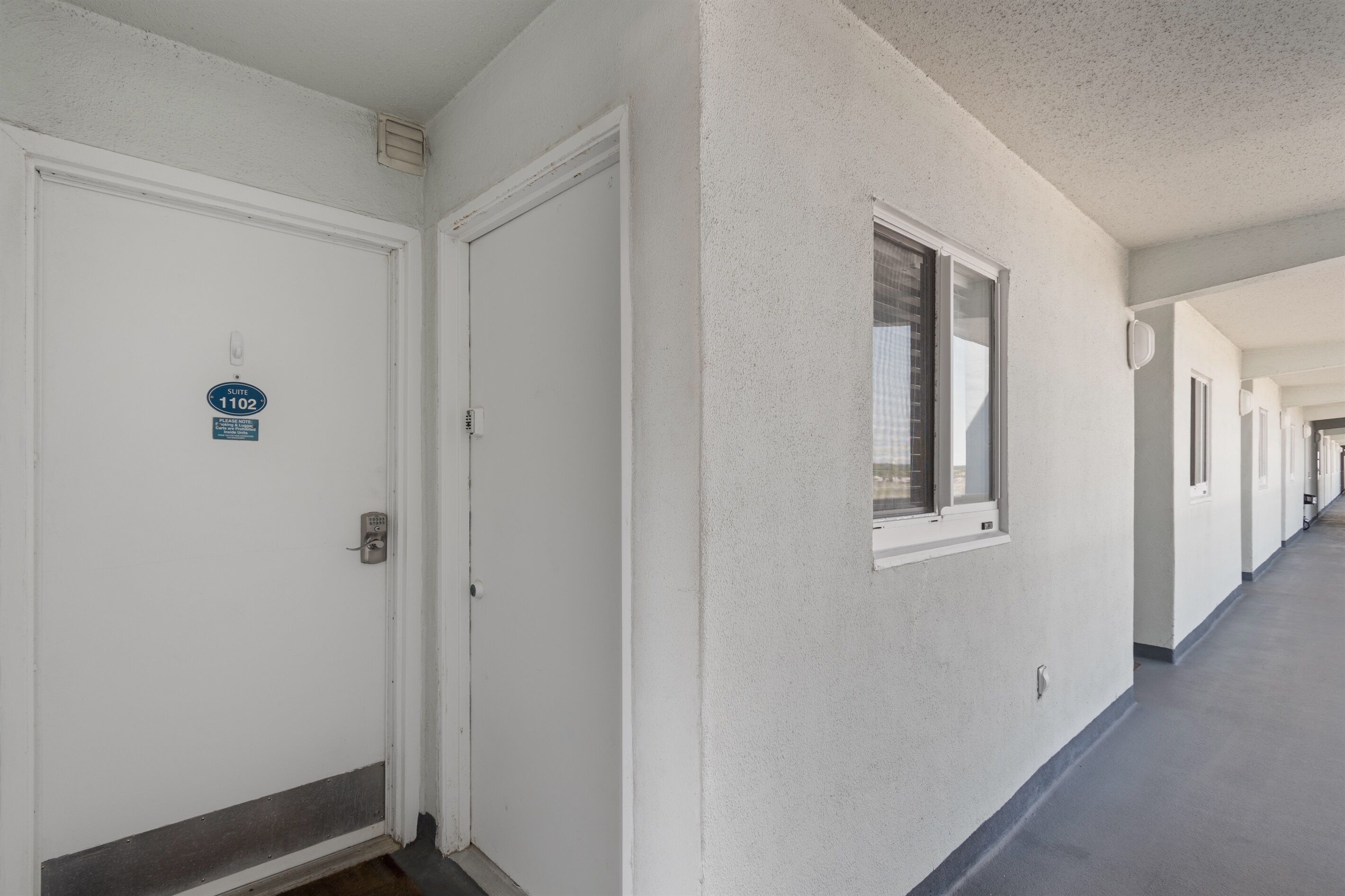 A long, narrow hotel corridor with white walls and doors, each marked with a number signifying room numbers.