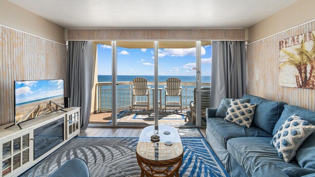 A balcony with a view of the ocean, a sofa, a TV, and a dining table. - Garden City Beach Oceanfront Condo 505 | Rising Tides Properties Rental (Murrells Inlet)