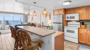 A kitchen with a large island, wooden cabinets, stainless steel appliances, wicker chairs, and a view to the outside. - Garden City Beach Oceanfront Condo 505 | Rising Tides Properties Rental (Murrells Inlet)