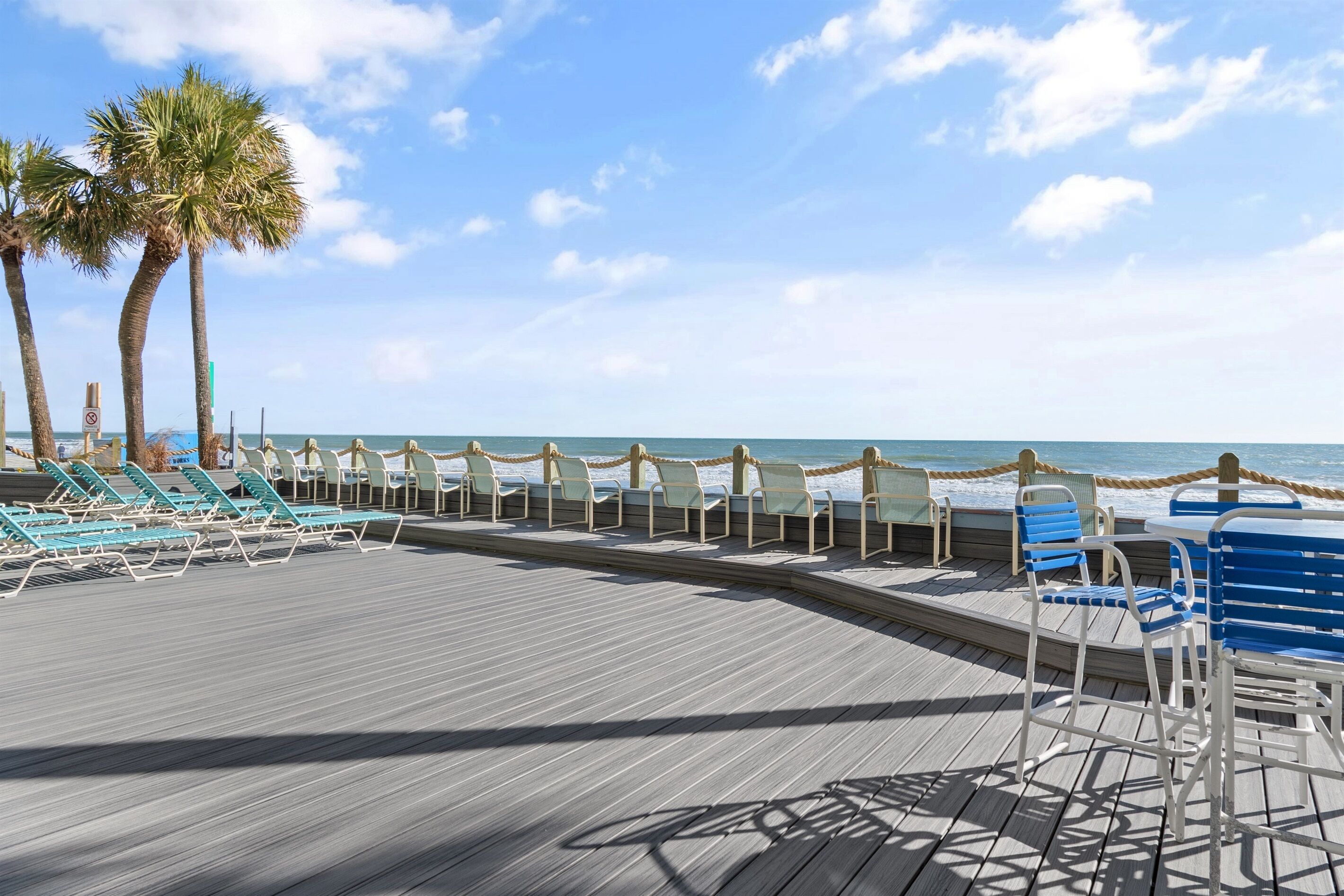 A wooden deck with lounge chairs and tables, palm trees, and a view of the ocean.