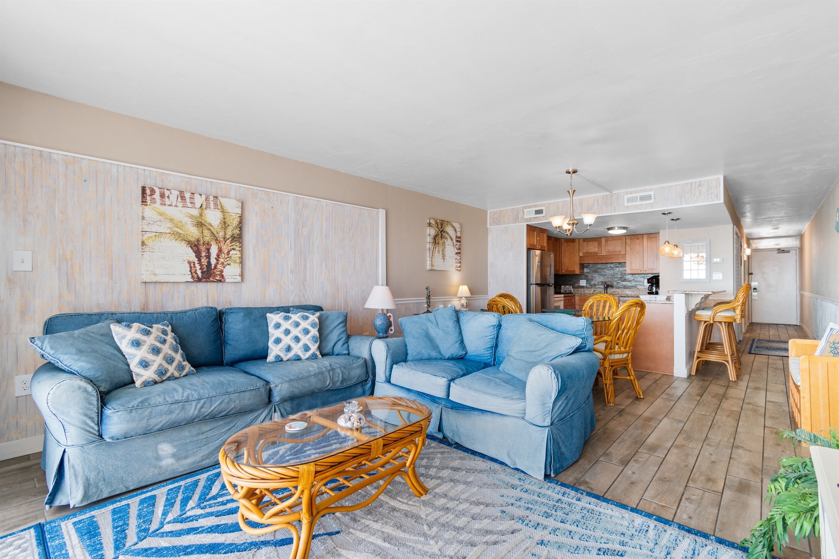 A living room with two blue sofas, a glass coffee table, and a dining area with wicker chairs.