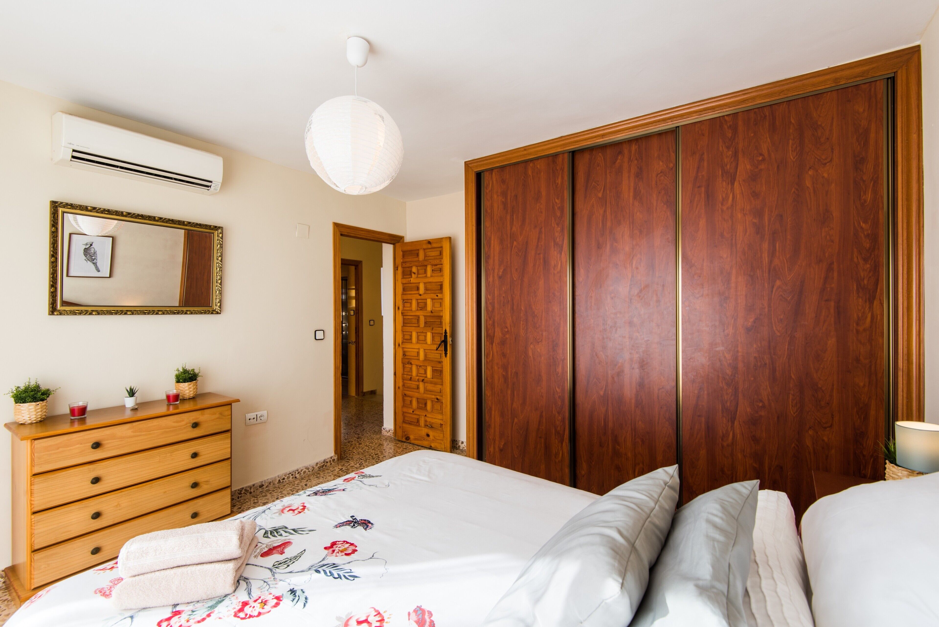 Une chambre avec une commode en bois, un lit avec une literie à motifs floraux, une armoire et un luminaire suspendu.