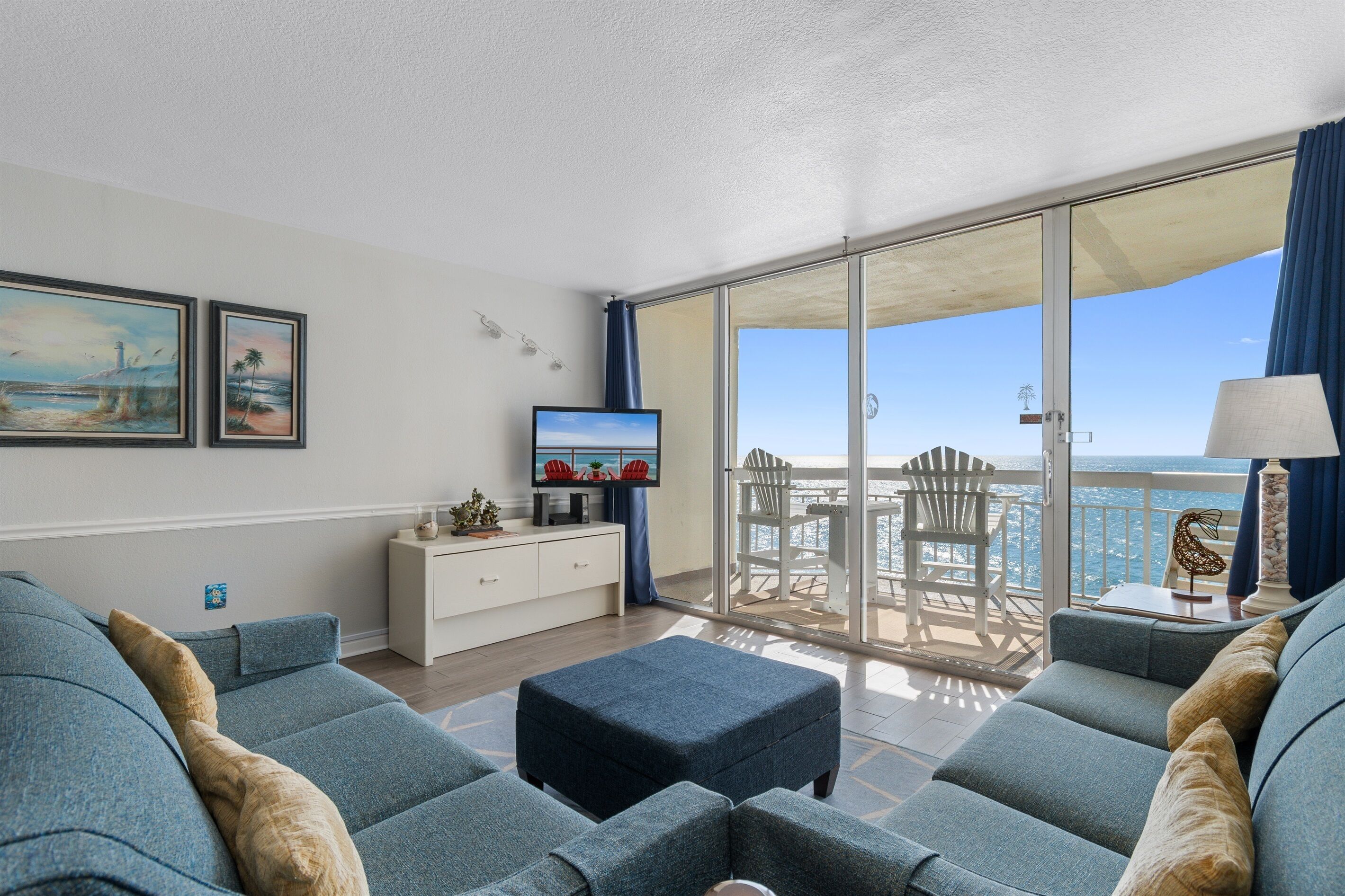 Un salon moderne avec un canapé, un pouf et un téléviseur à écran plat posé sur un meuble blanc. On aperçoit un balcon avec une table et des chaises, d’où l’on a une vue sur la mer.