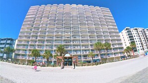 A large, multi-story hotel with numerous balconies, a beach in the foreground, and palm trees. - Garden City Beach Oceanfront Condo 910 | Rising Tides Properties Rental (Murrells Inlet)