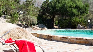 Area piscina con bordo in pietra, un asciugamano rosso su una sedia a sdraio e alberi circostanti.