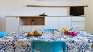Zona pranzo con tavolo apparecchiato per due, televisore sopra i pensili e mensola con oggetti decorativi.