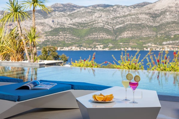 Área da piscina com vista para montanhas e mar. Há uma espreguiçadeira com uma revista, uma mesa com frutas e bebidas, e uma piscina com água cristalina.