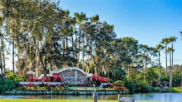 A sign for Heritage Oaks Golf & Country Club surrounded by lush greenery and a pond with two herons. - Heritage Oaks 2 Bedroom with Golf Course and Water Views (Sarasota)