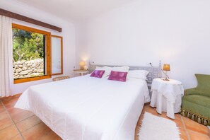 A bedroom with a large bed, a nightstand, a chair, a window with a view of greenery, and a stone wall.