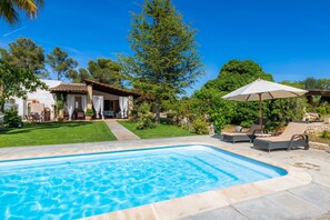 A swimming pool with lounge chairs and an umbrella, surrounded by greenery and a building with a tiled roof.