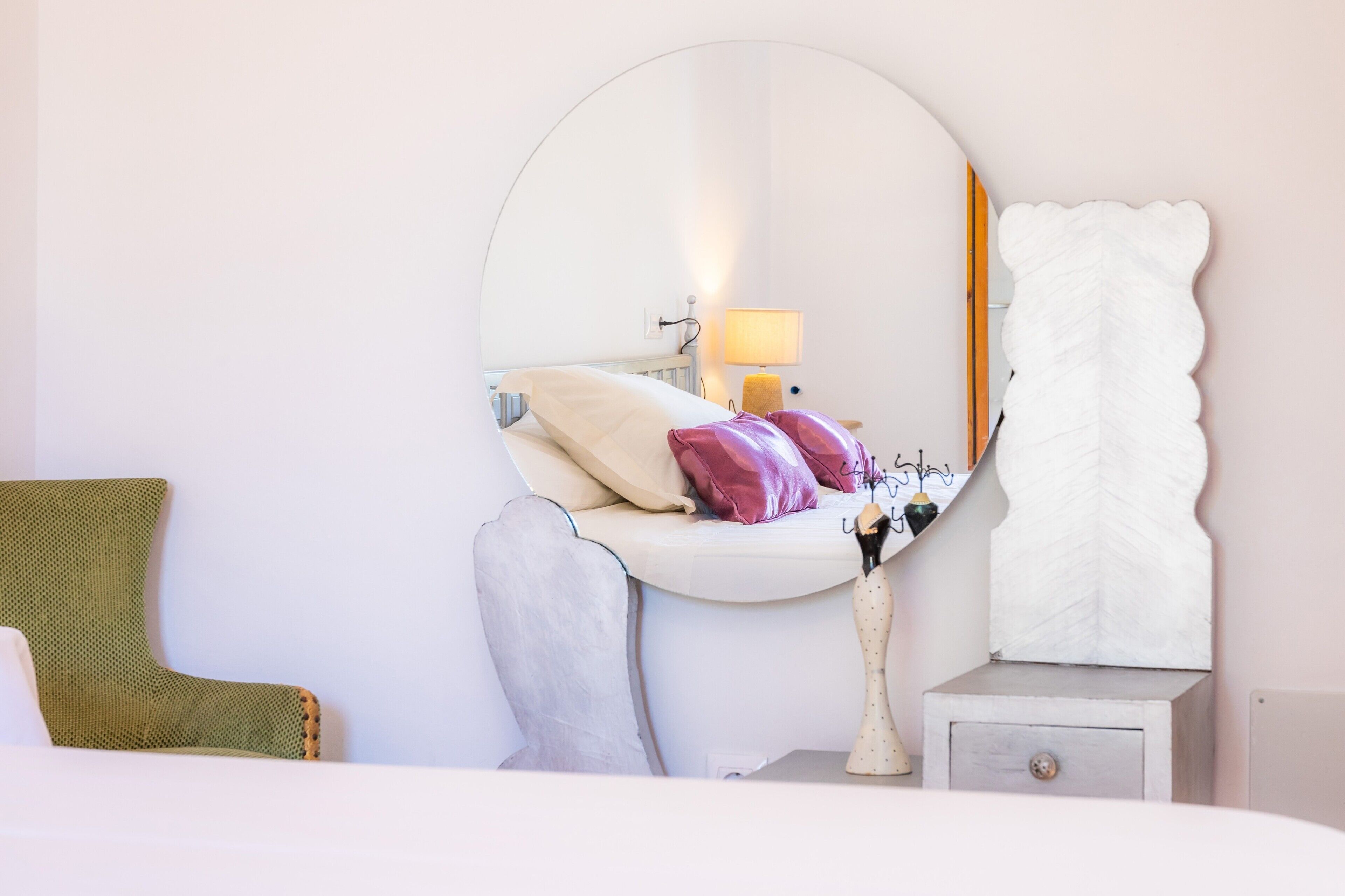 A bedroom with a round mirror, a bedside table with a lamp, a chair, and a bed with pillows.