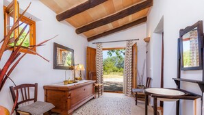 Habitación con escritorio de madera, una silla y una mesita con un jarrón. Se ve una ventana con cortinas, una puerta que da al exterior y un espejo en la pared.