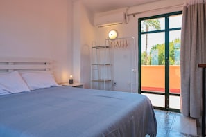 Un dormitorio con cama, mesita de noche, aire acondicionado instalado en la pared, ventana con vistas a zonas verdes y puerta con toallero.