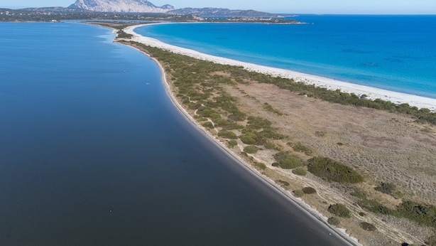 Vue aérienne d’une zone côtière avec une plage, des eaux d’un bleu limpide et une montagne en arrière-plan.