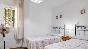 Ein Schlafzimmer mit zwei Betten, einem Standventilator, einem Fenster mit Vorhängen und einem kleinen Nachttisch mit Lampe.