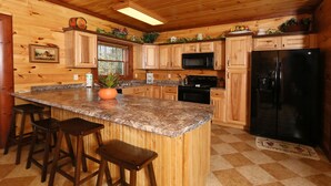 A wooden cabin kitchen with granite countertops, wooden cabinets, a black refrigerator, and a window overlooking the forest. - Splash N' Play #524 (Cosby)