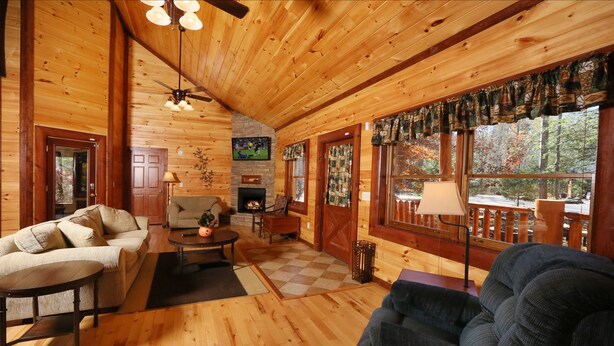 A cozy cabin interior with wooden walls, a fireplace, a ceiling fan, and large windows offering a view of the forest. - Splash N' Play #524 (Cosby)