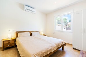 A bedroom with a bed, bedside tables, an air conditioner, a window, and a door.