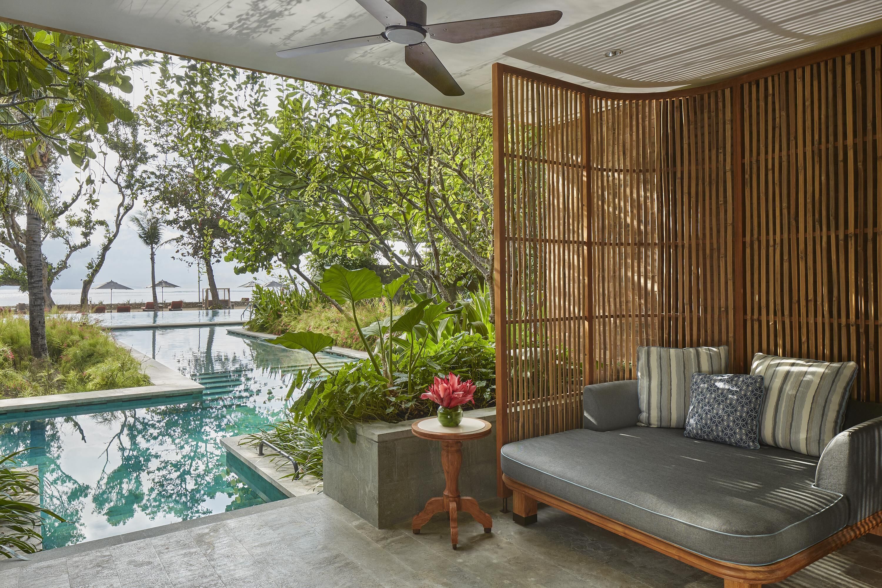 A modern outdoor seating area with a pool, surrounded by lush greenery and a view of the sea.