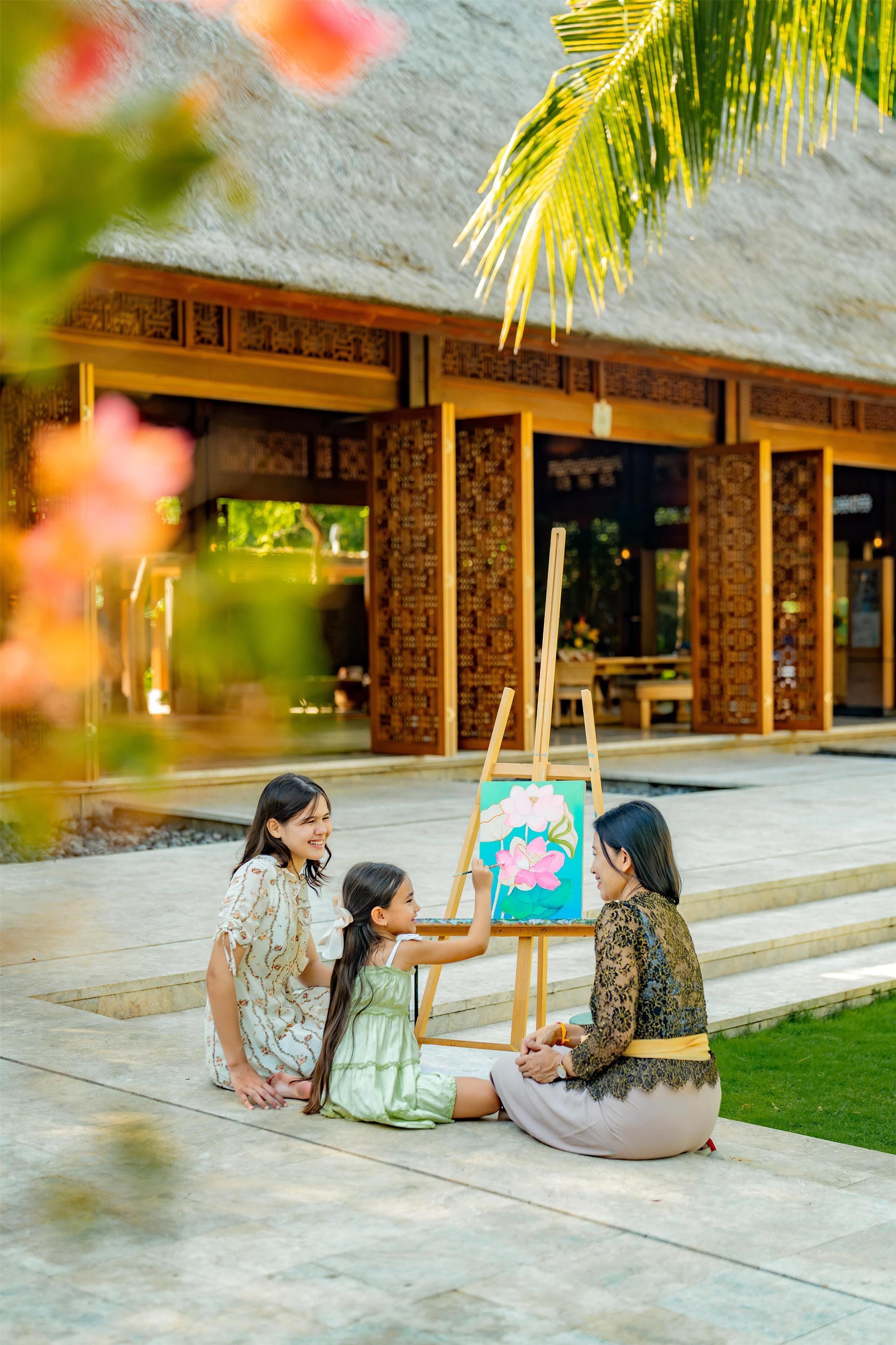 A group of people sitting on the ground in front of a building with a thatched roof and wooden columns.