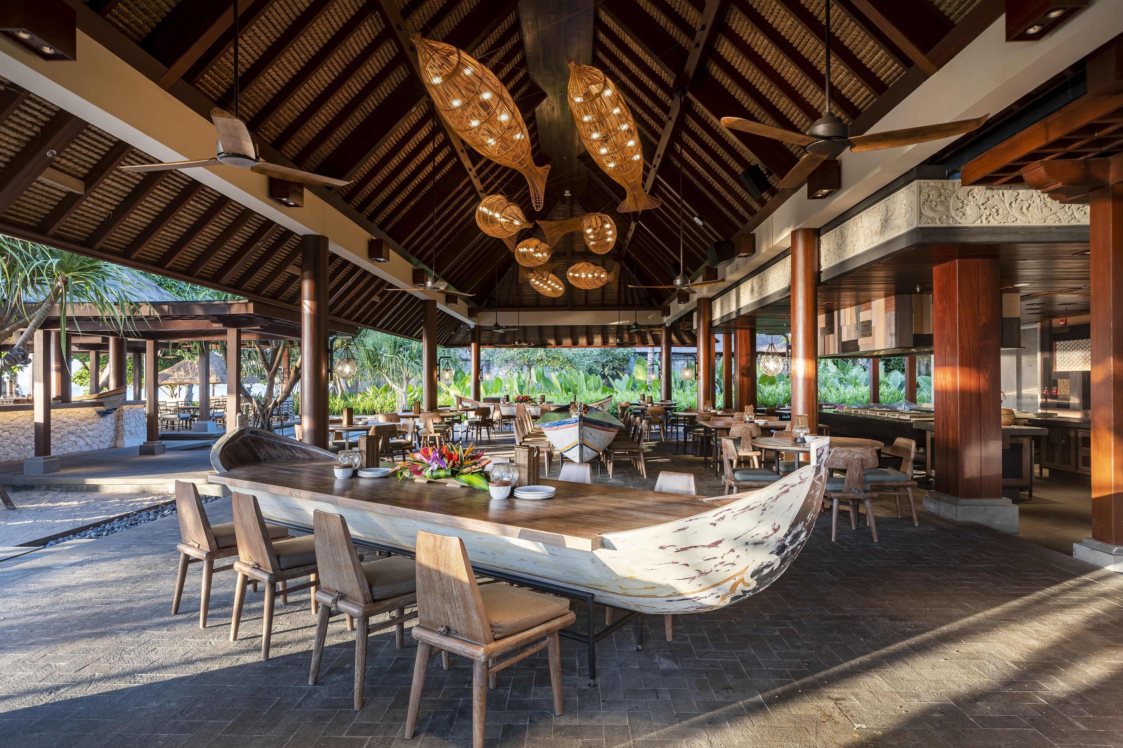 A spacious outdoor dining area with a long wooden table and chairs, surrounded by large windows and a view of greenery.