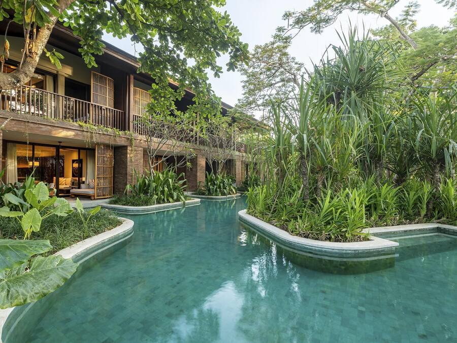 A pool area with a curved design, surrounded by lush greenery and a building with a balcony.
