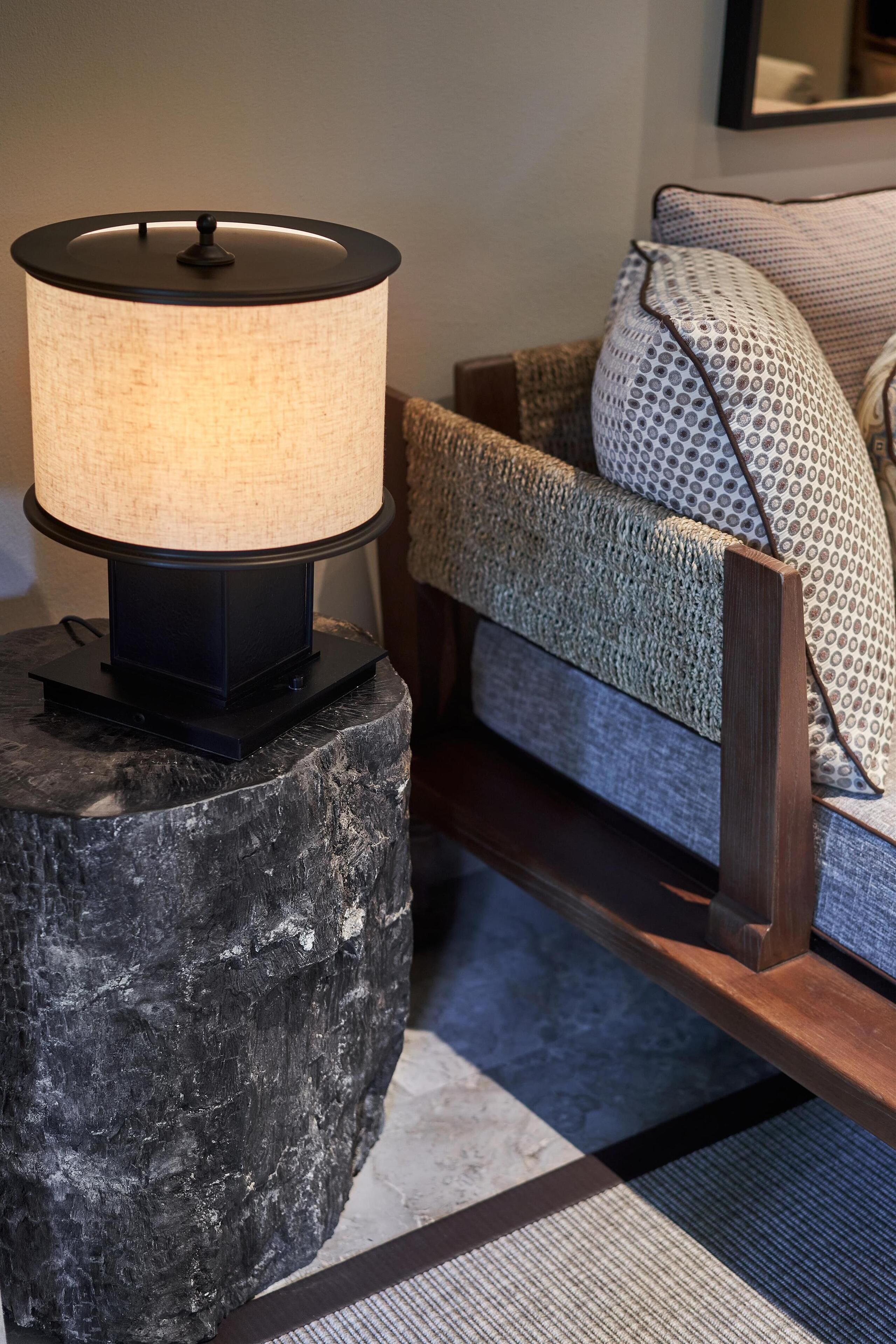 A lamp on a dark stone base, a wooden bench, and a patterned cushion on a couch.