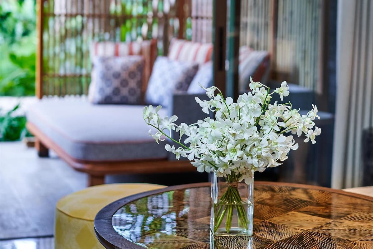 A room with a glass-top table, a vase with white flowers, a sofa with patterned cushions, and a bamboo screen in the background.