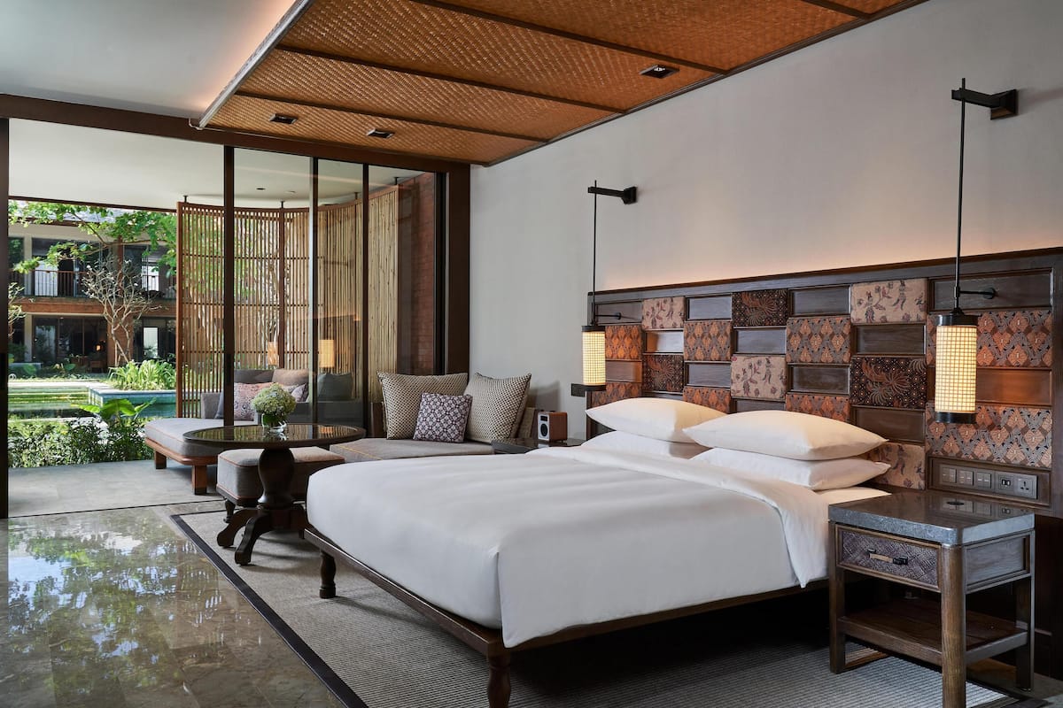 A modern hotel room with a large bed, a bedside table, a lamp, and a view of a garden and pool.