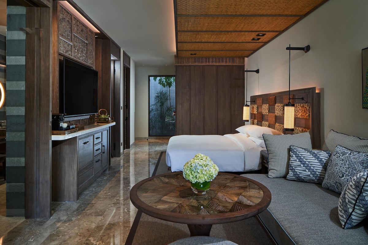 A modern hotel room with a large bed, a sofa, a dining area with a TV, and a decorative vase.