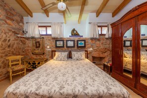 Une chambre avec un mur en pierre, des poutres apparentes en bois au plafond, un lit avec une literie à motifs floraux, une armoire en bois et une cheminée.