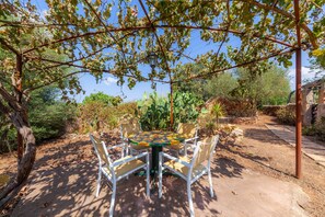 Une table ronde entourée de chaises, placée sous une pergola couverte de plantes grimpantes, le tout étant bordé d’un jardin où poussent différentes espèces végétales et d’un mur en pierre.