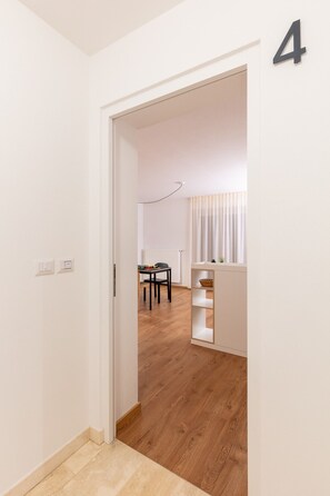 Un couloir mène à une pièce dotée d’un plancher en bois, d’une table avec des chaises, d’un radiateur et d’une petite plante posée sur une étagère.
