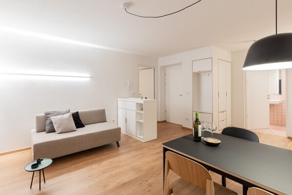 Une pièce moderne et bien éclairée, comprenant un canapé, une table à manger avec des chaises, une petite table d’appoint et un meuble avec un miroir.