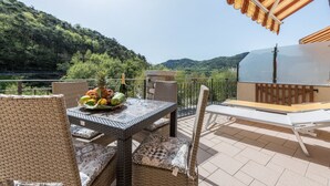 A patio with wicker furniture, a table with fruit and a bottle, and a view of a forested hill. - Beautiful Apartment “Residence Tatiana A3” with Pool, Terrace (Tremosine)