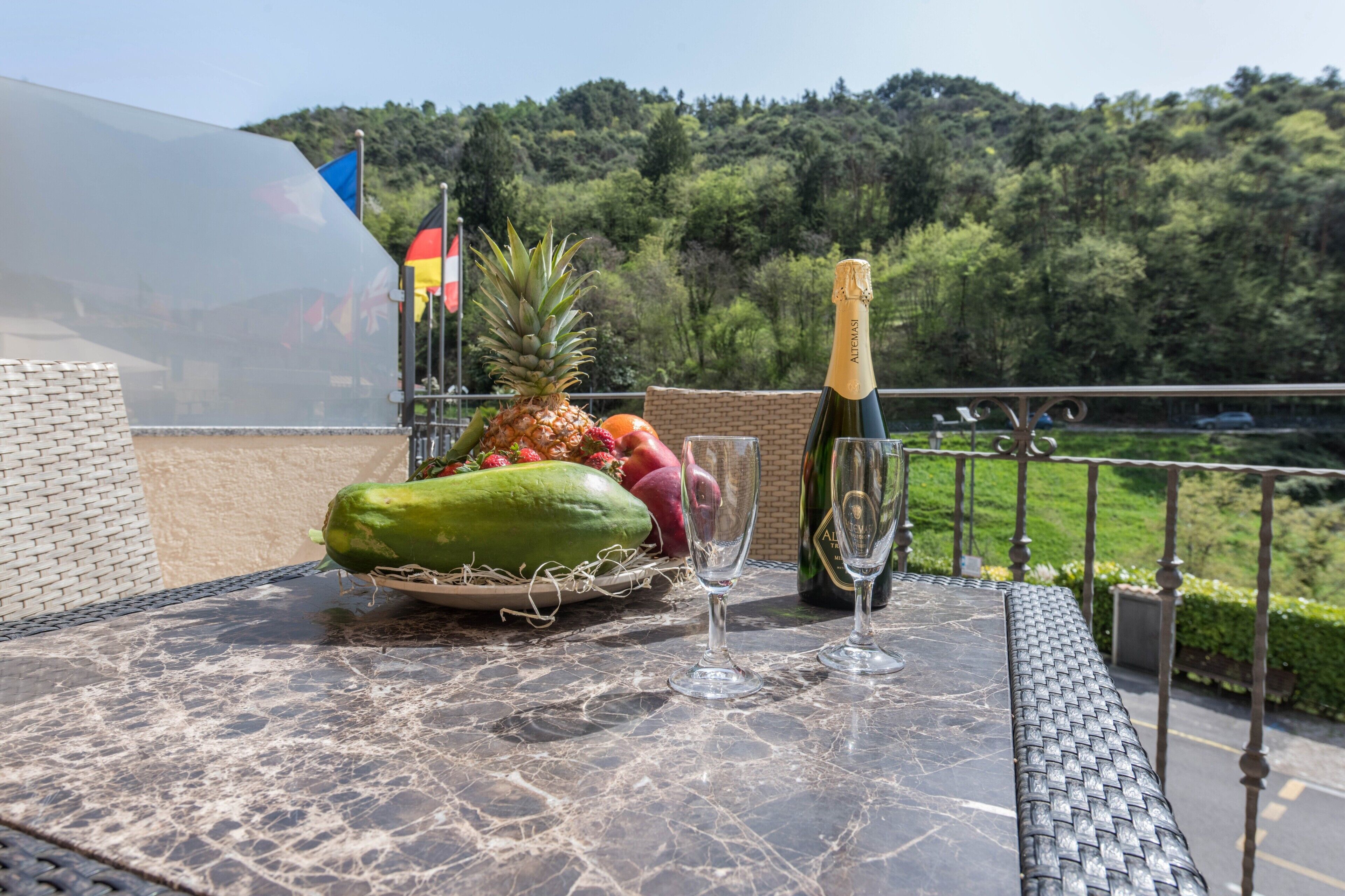 Une table avec une bouteille de champagne, des verres et un panier de fruits, placée sur un balcon donnant sur une zone boisée.