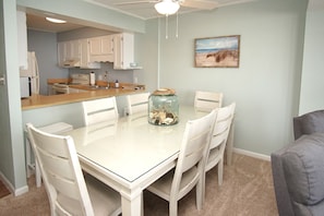Uma sala de jantar com uma mesa e cadeiras brancas, um pote de vidro com areia e conchas, e um quadro com tema praiano na parede.