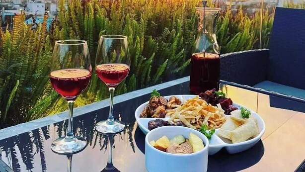 A table with a glass of red wine, a glass of white wine, and a glass of dark beverage, along with a plate of food and a bowl of fruit, set against a scenic view of a city and a sunset.