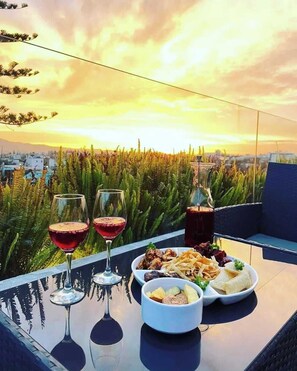 Een tafel met een glas rode wijn, een glas witte wijn en een glas donker drankje, plus een bord eten en een schaal fruit, met op de achtergrond een mooi stadsgezicht en een zonsondergang.