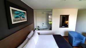 A hotel room with a bed, a blue armchair, a small table, a potted plant, and a large window with a view of a tree and a bathtub.