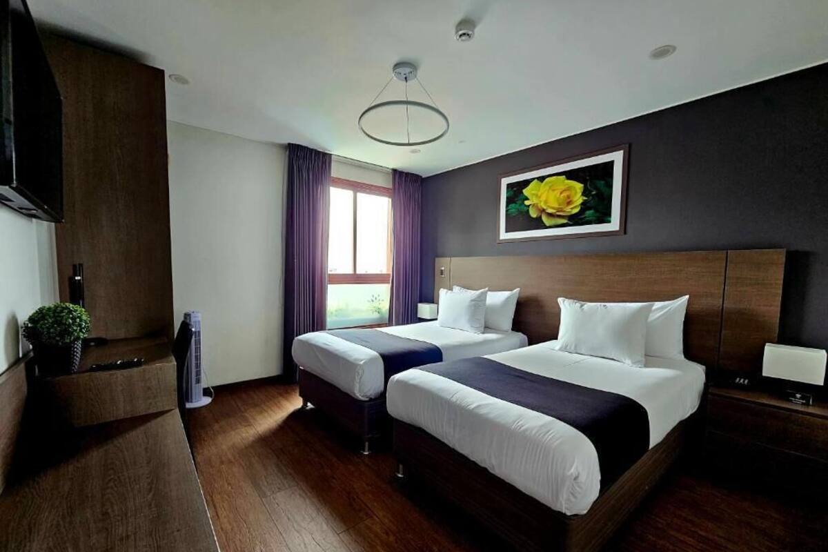 Een hotelkamer met twee bedden, een groot raam met gordijnen, een wandtelevisie en een ingelijst schilderij aan de muur.