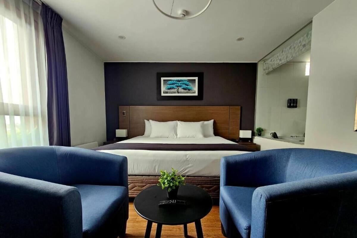 Een hotelkamer met een groot bed, twee blauwe fauteuils, een kleine ronde tafel met een plant en een schilderij aan de muur.
