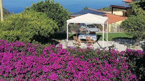 Un espace extérieur aménagé, abrité et doté de tables et de chaises, entouré de fleurs roses éclatantes, offrant une vue sur un paysage côtier avec la mer et des montagnes lointaines.