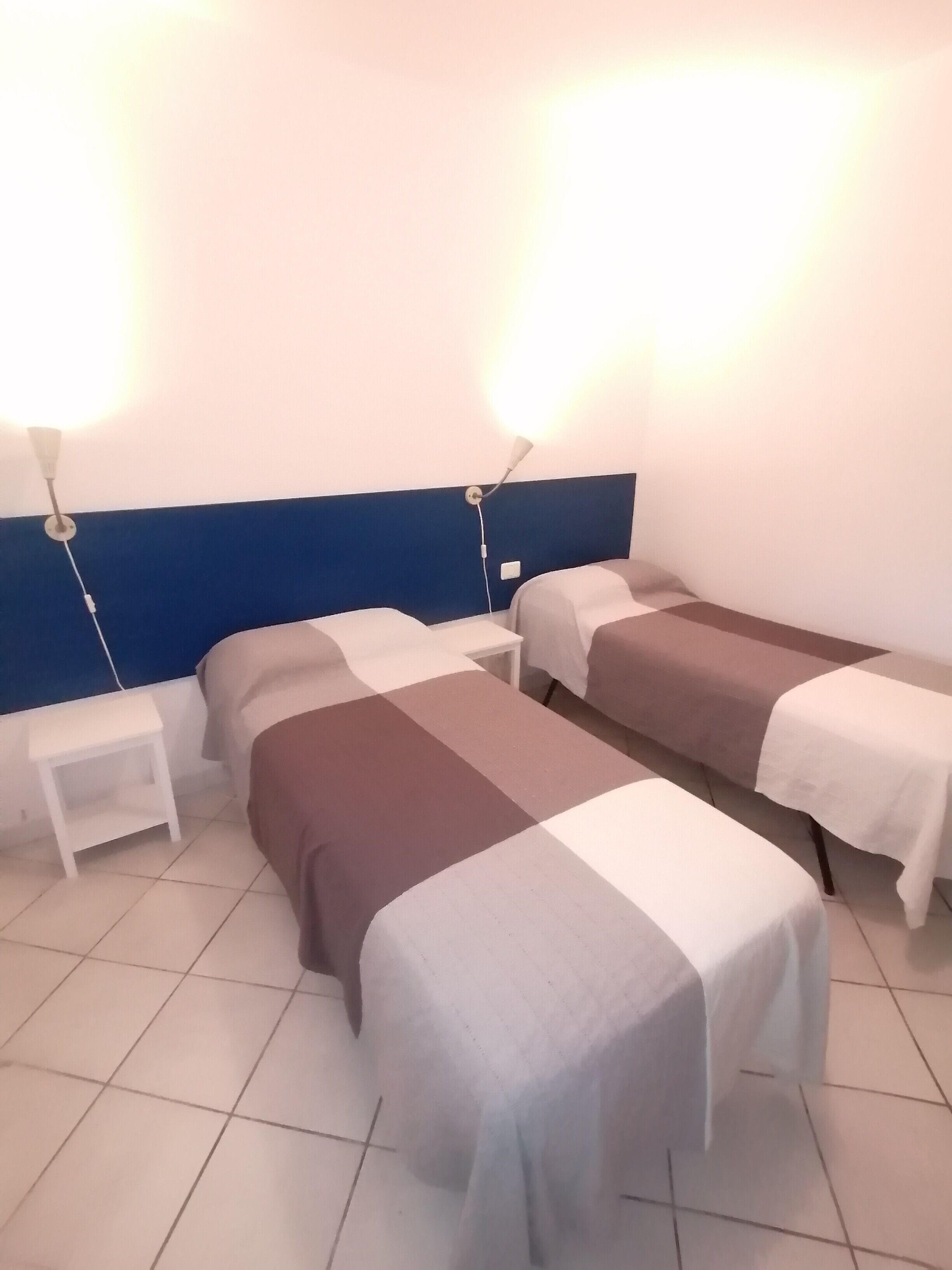 A hotel room with two beds, a nightstand, and a wall-mounted lamp.