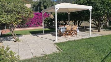 Een overdekt terras met een tafel en stoelen, omgeven door een verzorgd gazon en bloeiende struiken.