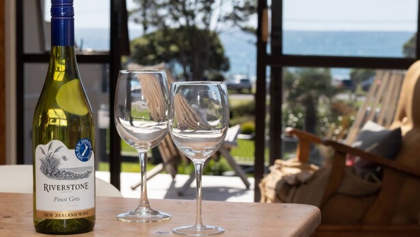 Een fles Riverstone Pinot Gris en twee glazen op een houten tafel, met uitzicht op zee en begroeiing.