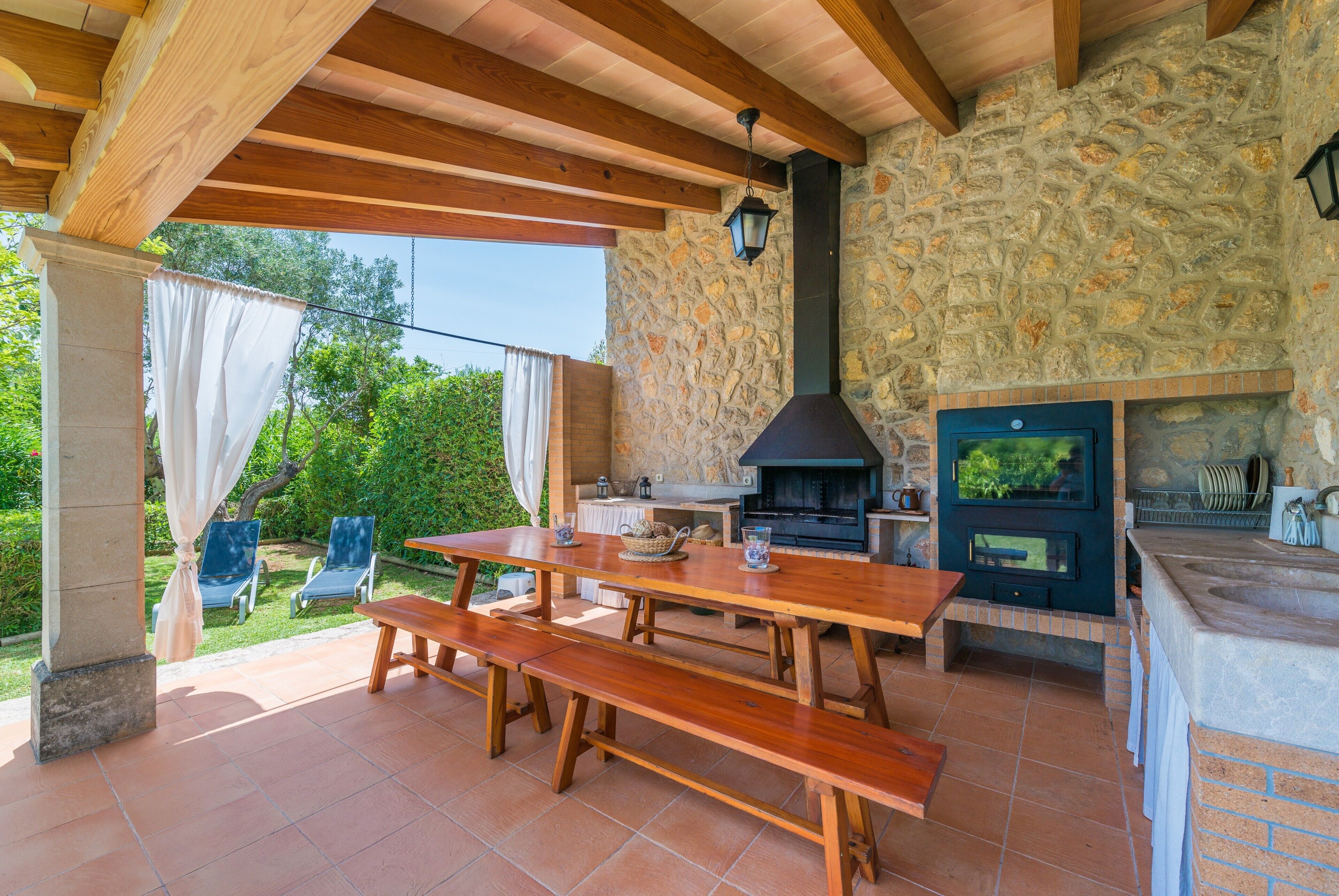 Un patio al aire libre con pared de piedra, vigas de madera, chimenea de piedra y una mesa con bancos.