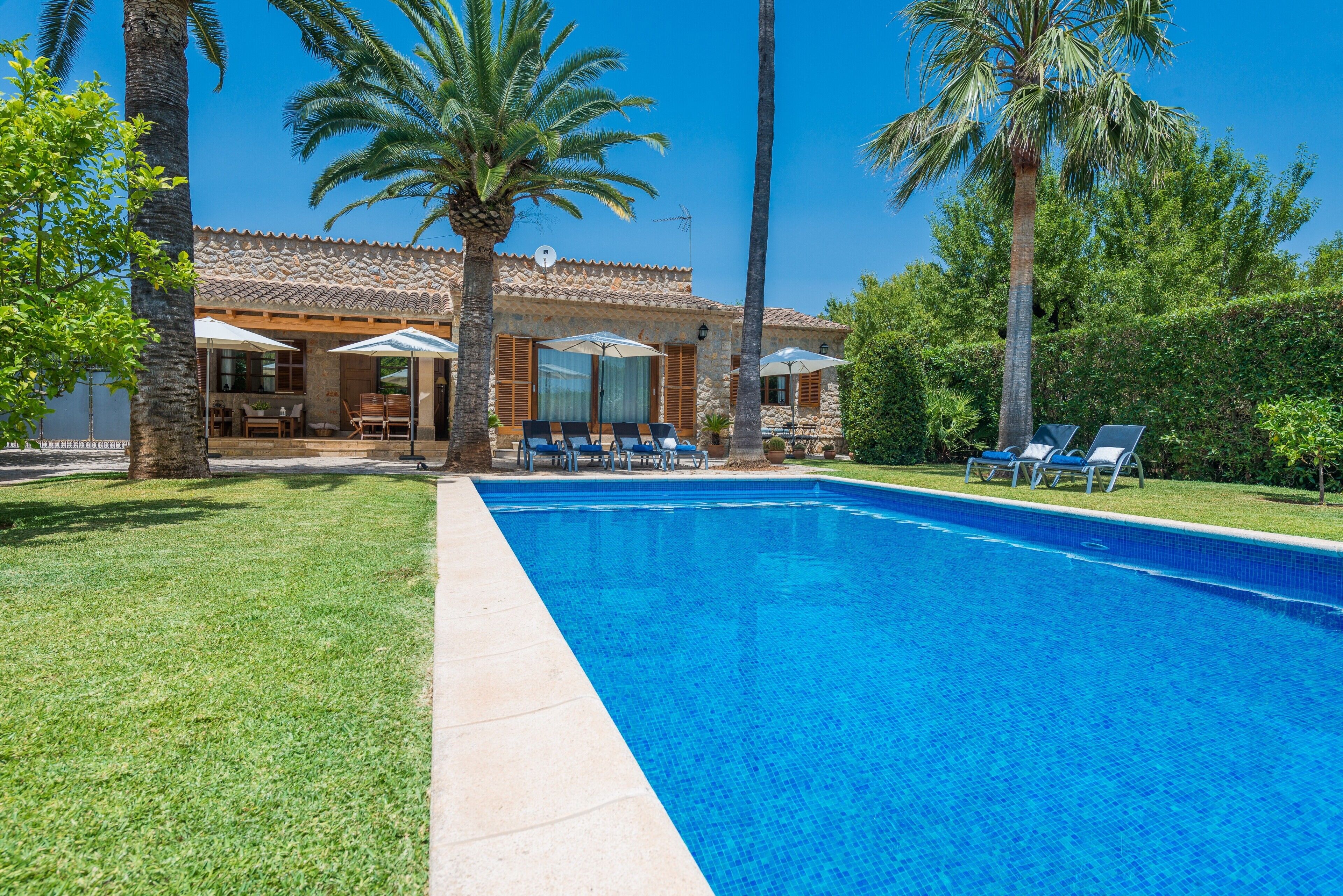 A swimming pool with lounge chairs, a patio area with umbrellas, and a stone building with a tiled roof.