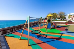 Un parque infantil con columpios y tobogán, suelo de colores y vista al mar.