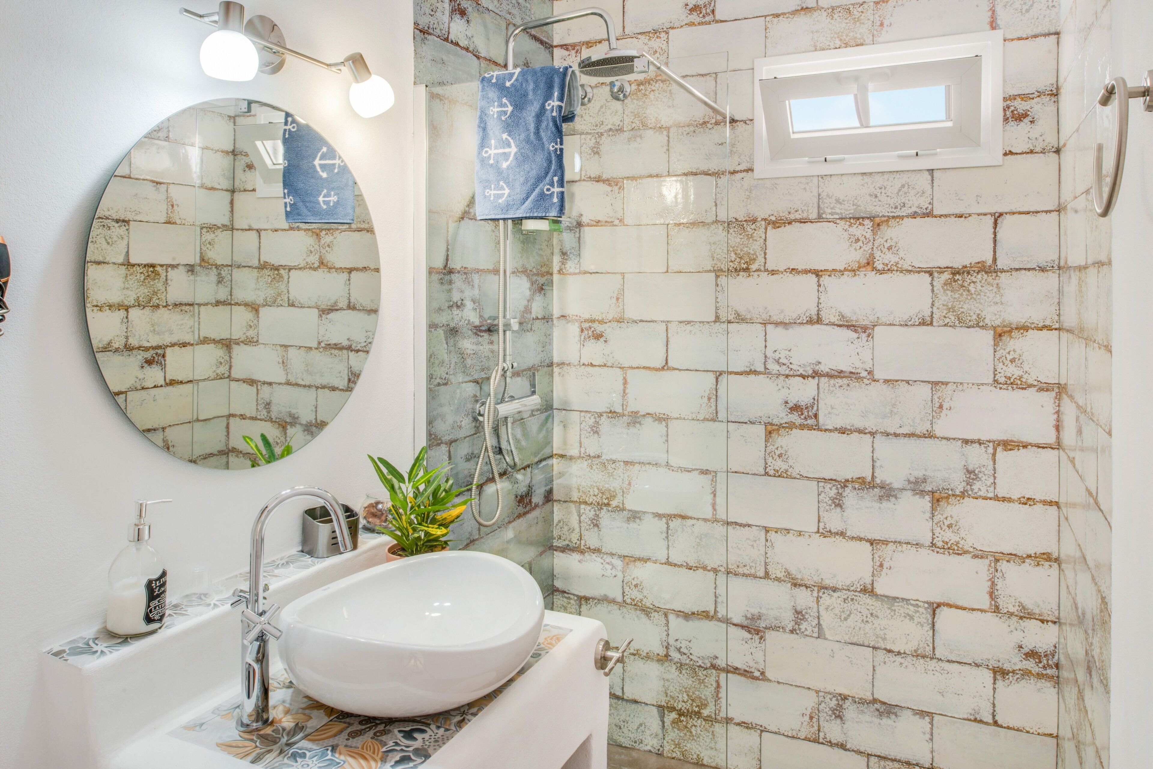 A bathroom with a round mirror, a sink, a shower, and a window.